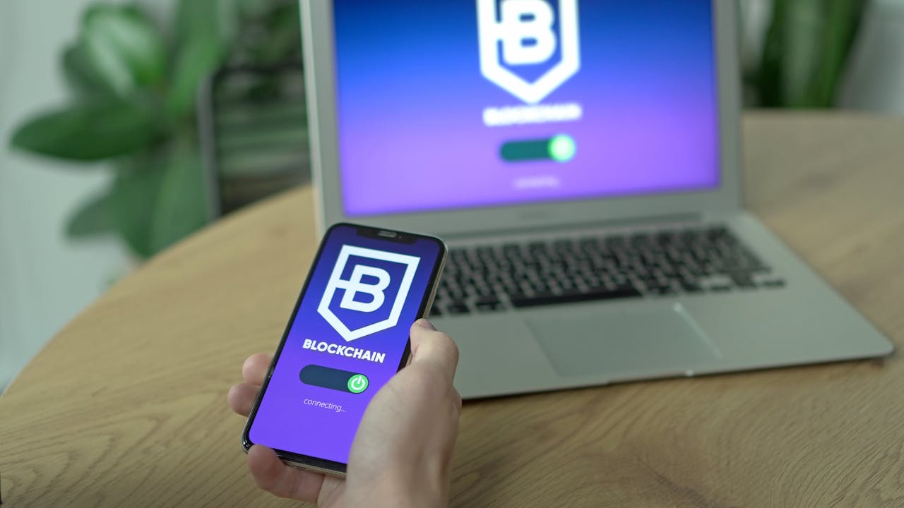 A hand holding a smartphone displaying a blockchain app, with a laptop on a desk in the background.